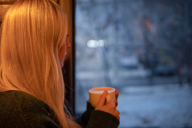 Vrouw met een kop koffie kijkt uit het raam op een besneeuwde dag. Ze denkt aan haar ex en is nog niet verdergegaan na de breuk.