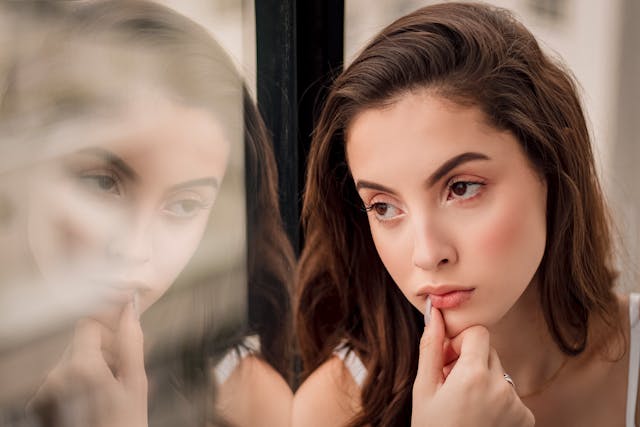 Vrouw kijkt peinzend uit het raam en ziet haar eigen weerspiegeling. Ze twijfelt of het wel de juiste beslissing was om de relatie te verbreken.