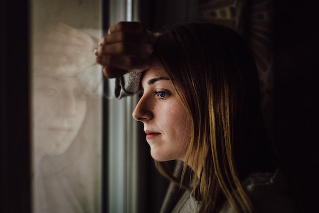 Vrouw kijkt peinzend uit het raam, diep in gedachten of ze haar ex moet geven wat hij wil, of juist niet.